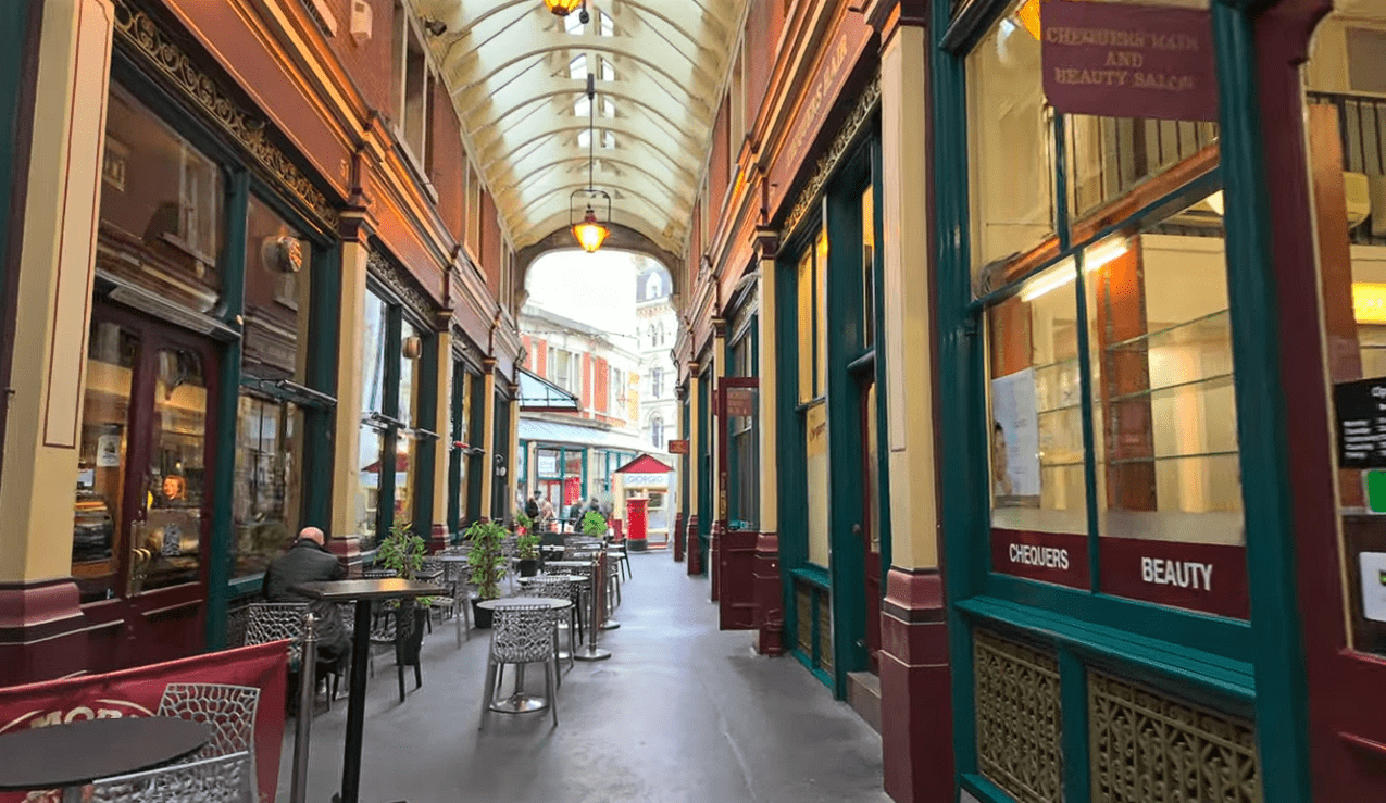 Interior walkway of Leadenhall Market with covered glass roof, shopfronts on both sides, and outdoor café seating.