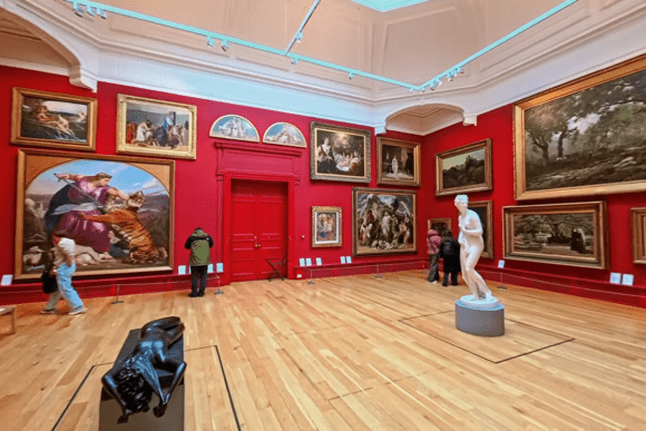 Grand exhibition room at Leeds Art Gallery featuring large classical paintings on striking red walls, elegant sculptures, polished wooden floors, and visitors exploring the historic art collection.