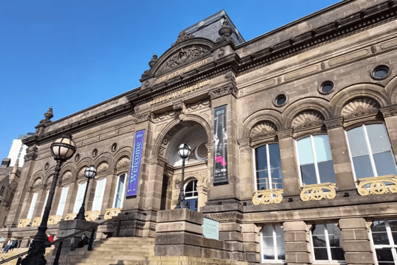 Historic exterior of Leeds City Museum in Leeds city centre, featuring grand Victorian architecture, stone columns, arched windows, and the main entrance beneath a clear blue sky.