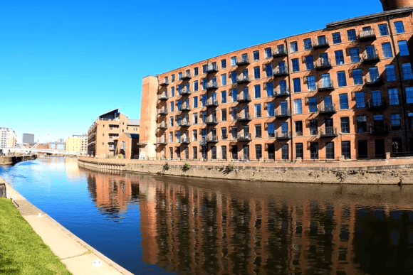 Historic red-brick riverside warehouse apartments in Leeds South Bank reflected in the River Aire under a clear blue sky.