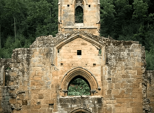 Stone façade of Mount Grace Priory with arched doorway and tower set against wooded hillside.