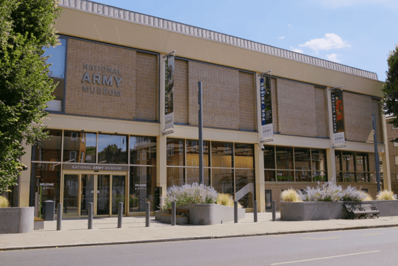 Front exterior of the National Army Museum in Chelsea, London, featuring a modern brick façade, large glass entrance, museum banners, landscaped planters, and a welcoming public plaza.