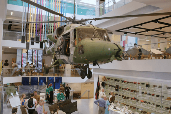 Military helicopter suspended inside the National Army Museum in London, displayed above visitors exploring interactive exhibits and collections showcasing the history of the British Army.