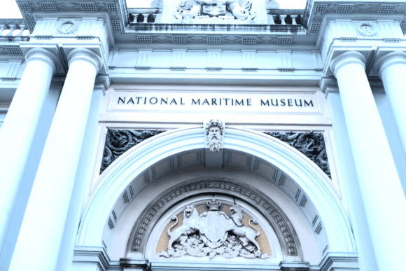 Exterior view of the National Maritime Museum in Greenwich, London, featuring a grand neoclassical entrance with towering white columns, ornate stone carvings, and the museum’s name displayed above the central archway.
