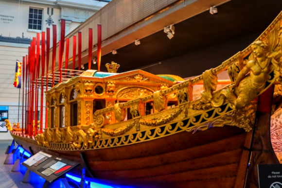Ornate royal barge displayed inside the National Maritime Museum in Greenwich, featuring gilded carvings, decorative figureheads, and vibrant red oars that highlight Britain’s rich maritime heritage.