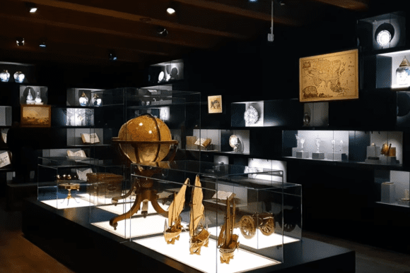 Gallery display inside the National Maritime Museum featuring a historic globe, antique maps, model sailing ships, navigation instruments, and maritime artefacts showcased in illuminated glass cases.