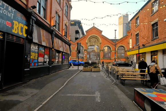 Street view in Manchester’s Northern Quarter with historic red-brick architecture, outdoor seating, colourful artwork and festoon lights.