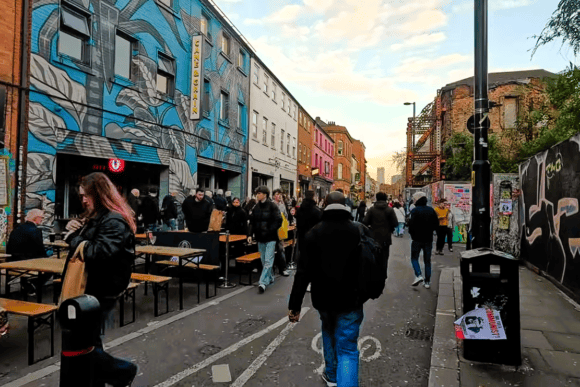 Busy street in Manchester’s Northern Quarter with colourful street art, outdoor seating, independent venues and pedestrians exploring the area.
