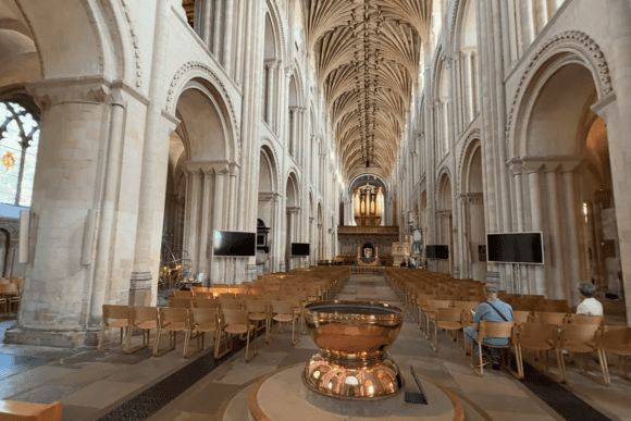 Interior of Norwich Cathedral nave with high vaulted ceiling, rows of wooden chairs, and a baptismal font in the foreground.