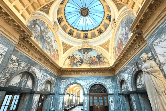 Interior of the Old Bailey featuring a domed ceiling with stained glass, painted murals, marble walls, and a statue overlooking an arched hallway.