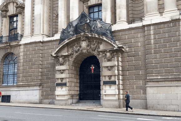 Entrance façade of the Old Bailey in London, featuring an ornate stone archway with sculptural detailing and a gated doorway, with pedestrians walking past.