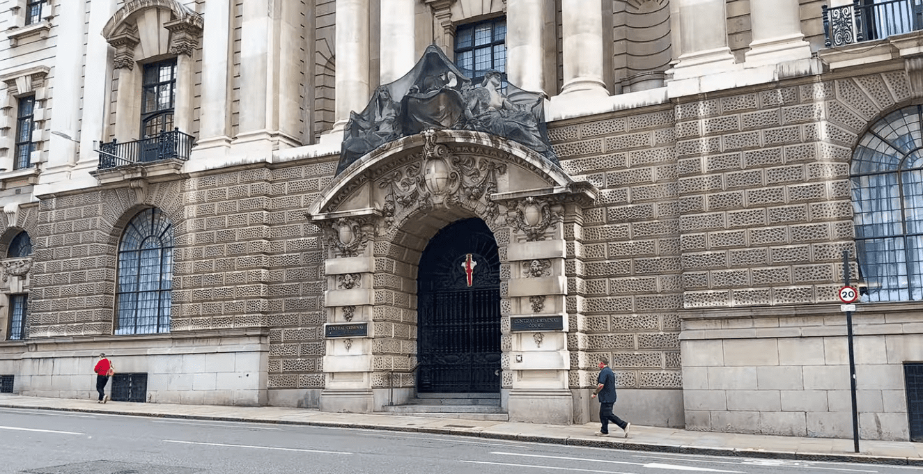 Entrance façade of the Old Bailey in London, featuring an ornate stone archway with sculptural detailing and a gated doorway, with pedestrians walking past.