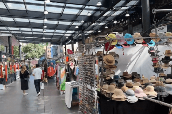 Market stall at Old Spitalfields Market displaying hats and sunglasses, with shoppers walking along a covered walkway.