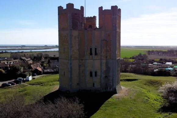 Aerial view of Orford Castle standing on a grassy mound near the coastline with surrounding village and fields.