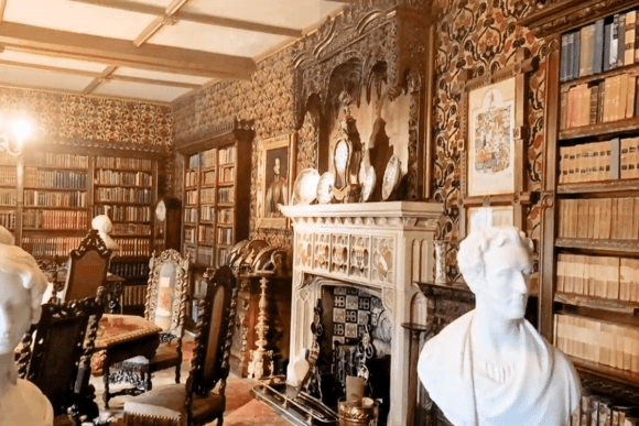 Historic library room at Oxburgh Hall with bookshelves, ornate fireplace, carved furniture, and classical busts.