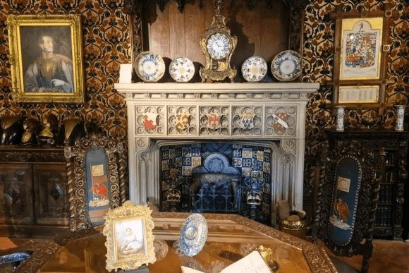 Ornate fireplace at Oxburgh Hall with decorative tiles, clock, porcelain plates, carved chairs, and framed artworks.