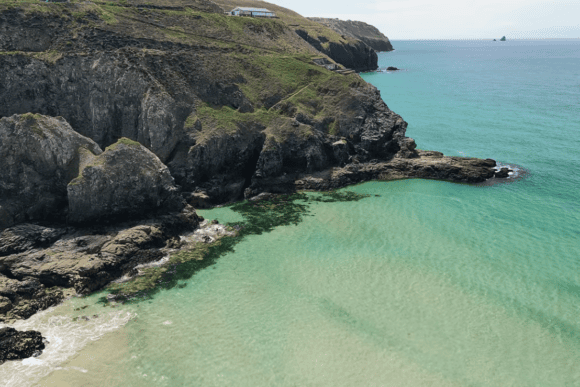 Aerial view of Perranporth Beach in Cornwall with crystal-clear turquoise water, rugged cliffs, and a secluded rocky cove along the Atlantic coastline.