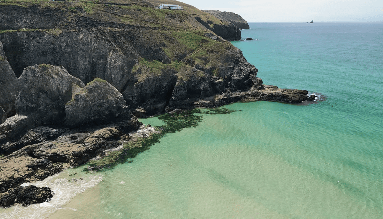 Aerial view of Perranporth Beach in Cornwall with crystal-clear turquoise water, rugged cliffs, and a secluded rocky cove along the Atlantic coastline.