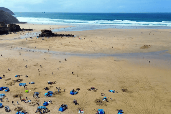 Aerial view of Perranporth Beach in Cornwall showing expansive golden sands, Atlantic waves, beachgoers, and dramatic coastal cliffs on a sunny day.