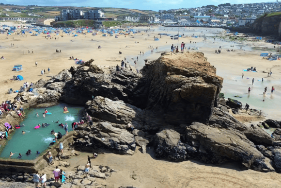 Aerial view of Perranporth Beach in Cornwall featuring the famous Chapel Rock tidal pool, golden sandy beach, beachgoers, and dramatic coastal scenery on a sunny summer day.