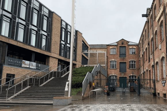 The Piece Hall in Halifax, West Yorkshire, showing historic red-brick buildings alongside modern architecture, with wet cobblestone paving and entrance steps on a rainy day.