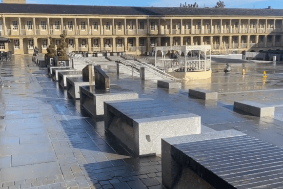 The Piece Hall courtyard in Halifax, West Yorkshire, featuring stone paving, seating benches, a central bandstand, and Georgian colonnades under clear skies after rain.