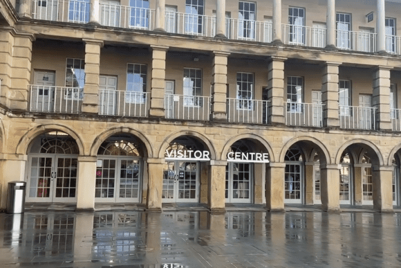 The Piece Hall Visitor Centre in Halifax, West Yorkshire, featuring arched stone colonnades, Georgian architecture, and wet reflective paving after rain.