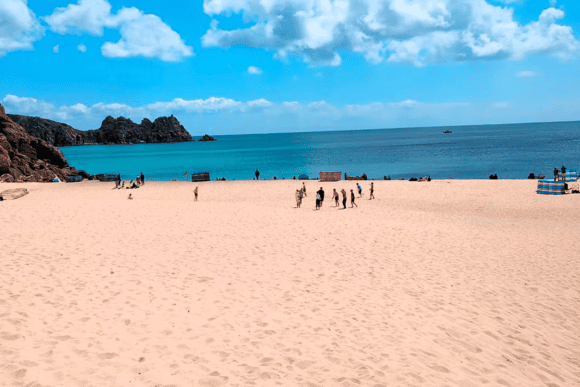 Porthcurno Beach with soft golden sand, crystal-clear turquoise sea, rugged granite rocks, and visitors enjoying one of Cornwall's most beautiful beaches under a sunny blue sky.