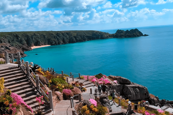 Elevated view of Porthcurno Beach with crystal-clear turquoise waters, rugged granite cliffs, vibrant coastal gardens, and scenic pathways overlooking Cornwall's dramatic coastline.