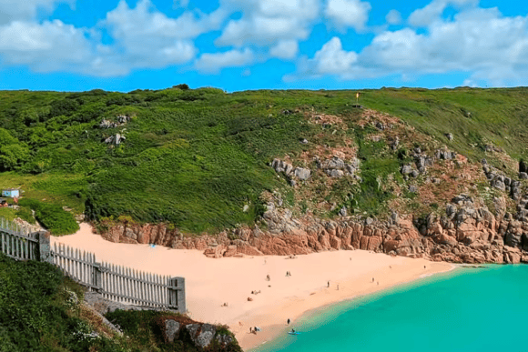 Porthcurno Beach with golden sand, turquoise waters, rugged granite cliffs, and lush green hills on Cornwall's dramatic coastline under a bright blue sky.