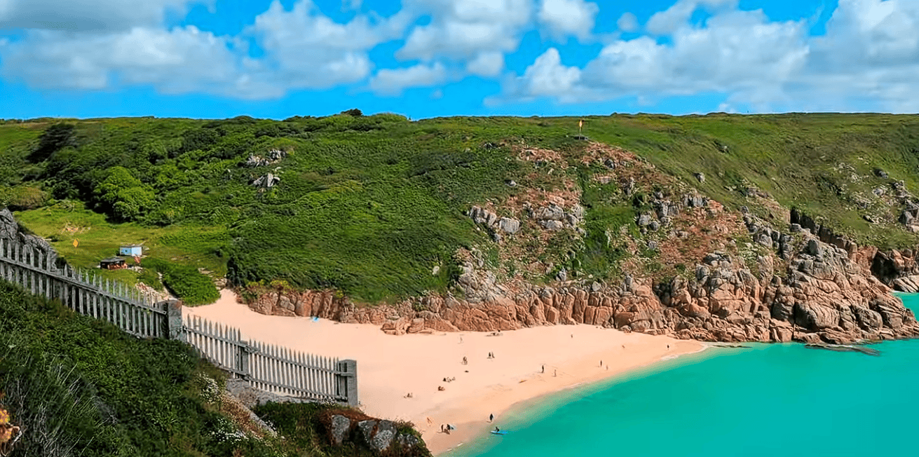 Porthcurno Beach with golden sand, turquoise waters, rugged granite cliffs, and lush green hills on Cornwall's dramatic coastline under a bright blue sky.
