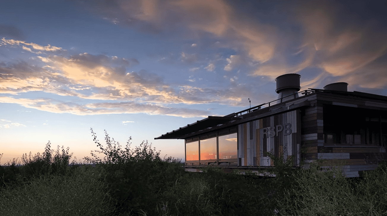 RSPB Rainham Marshes Visitor Centre at sunset, with dramatic clouds reflecting in the building’s glass façade, surrounded by wetlands and nature reserve vegetation in Essex.