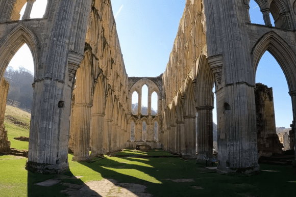 Interior ruins of Rievaulx Abbey in North Yorkshire, England, featuring tall Gothic arches, stone columns, and open roof with sunlight.