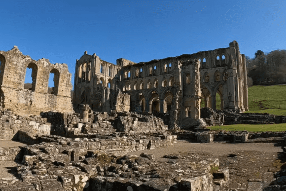 Rievaulx Abbey ruins in North Yorkshire, England, featuring extensive stone remains, Gothic arches, and open grounds under clear blue skies.