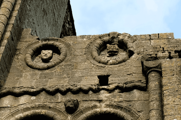 Close-up of carved stone faces and Romanesque decorative details on the exterior wall of Castle Rising Castle in Norfolk, England.