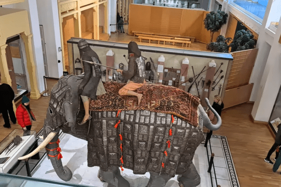 War elephant armour display at the Royal Armouries Museum in Leeds, featuring a life-sized armoured elephant model with mounted riders, surrounded by historic weapons and military artefacts.