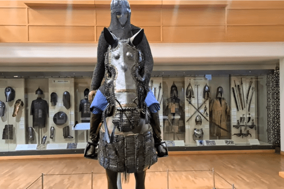 Front-facing display of a fully armoured medieval knight on horseback at the Royal Armouries Museum in Leeds, showcasing chainmail, horse armour, helmets, swords, and historic weapons in a military history gallery.