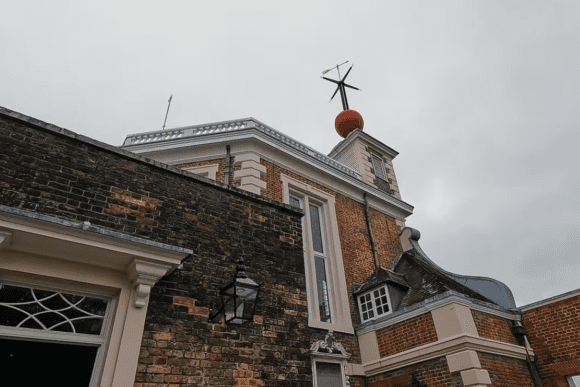 Close-up of Royal Observatory Greenwich building with red time ball and weather vane against a cloudy sky in London.