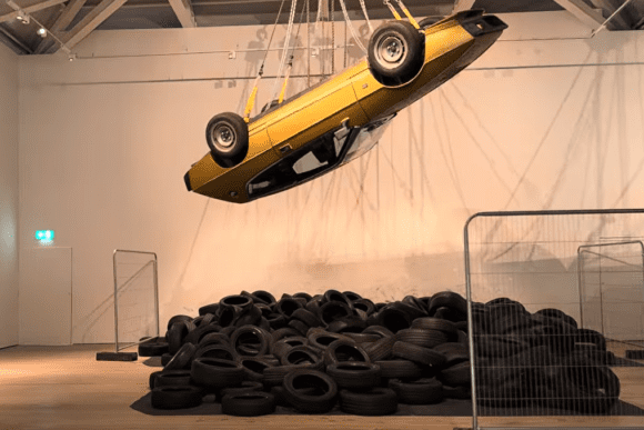 Suspended gold car installation above a large pile of black tyres at Saatchi Gallery in London, creating a dramatic contemporary artwork exploring consumer culture, transport, and urban environments.