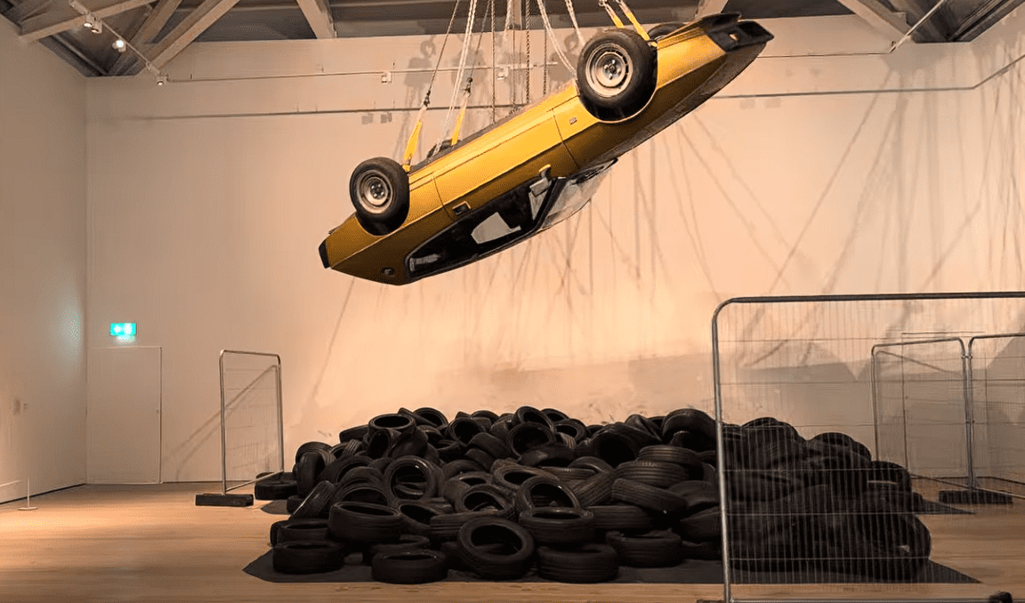 Suspended gold car installation above a large pile of black tyres at Saatchi Gallery in London, creating a dramatic contemporary artwork exploring consumer culture, transport, and urban environments.