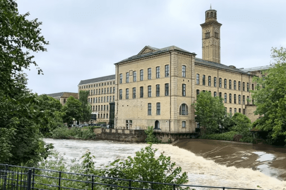 Salts Mill building beside the River Aire in Saltaire, West Yorkshire, with flowing water and surrounding greenery.