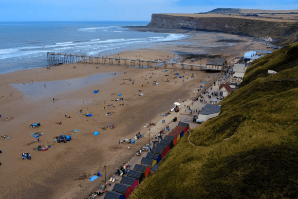 Aerial view of Saltburn Beach in North Yorkshire featuring the historic Saltburn Pier, golden sandy beach, colourful beach huts, dramatic cliffs, and the scenic Yorkshire coastline.