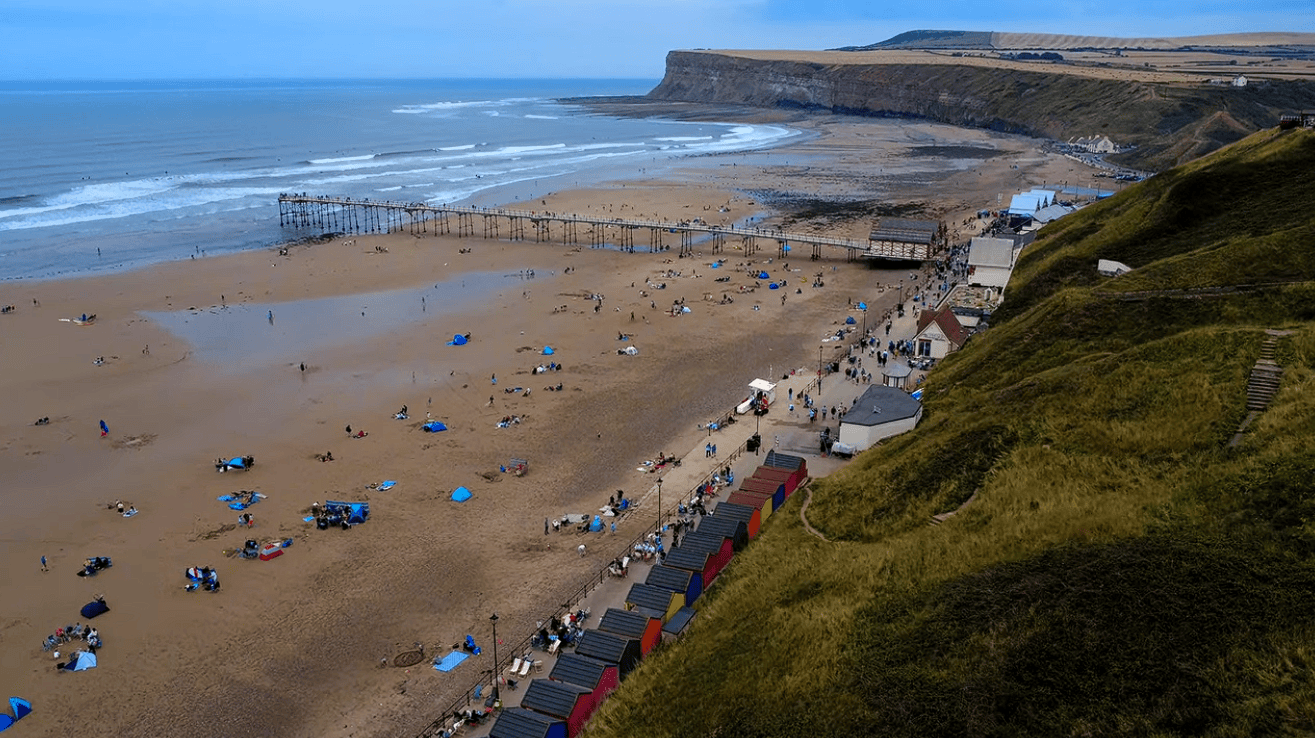 Aerial view of Saltburn Beach in North Yorkshire featuring the historic Saltburn Pier, golden sandy beach, colourful beach huts, dramatic cliffs, and the scenic Yorkshire coastline.