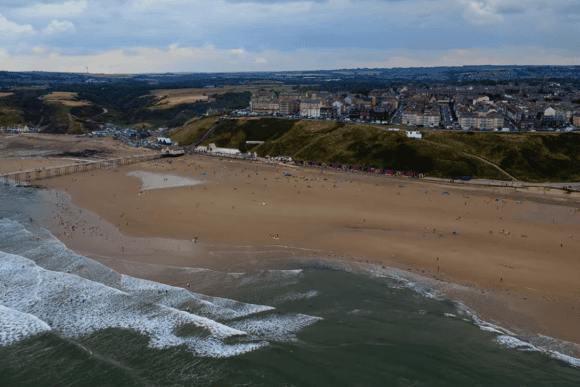 Aerial view of Saltburn Beach in North Yorkshire, showing golden sands, rolling waves, the historic Saltburn Pier, colourful beach huts, and the seaside town along the dramatic Yorkshire coastline.