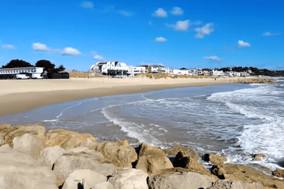 Wide view of Sandbanks Beach in Dorset with golden sand, gentle waves, rocky shoreline, and luxury beachfront homes under a clear blue sky.