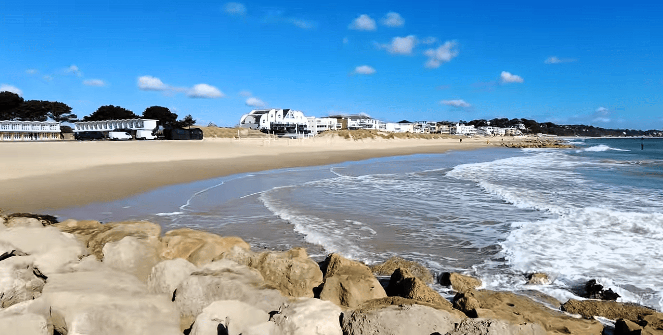 Wide view of Sandbanks Beach in Dorset with golden sand, gentle waves, rocky shoreline, and luxury beachfront homes under a clear blue sky.