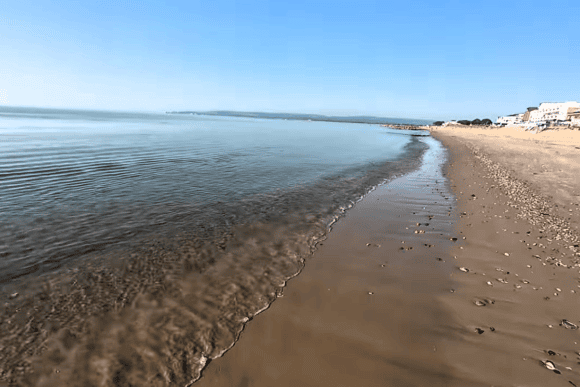 Peaceful shoreline at Sandbanks Beach in Dorset with calm, clear water, smooth wet sand, and luxury beachfront properties under a bright blue sky.