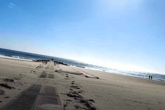 Accessible boardwalk leading across the golden sands of Sandbanks Beach in Dorset toward the sea on a bright, sunny day with sparkling blue water.