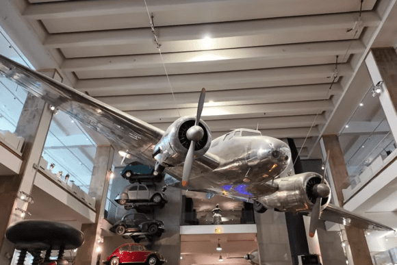 Historic silver aircraft suspended inside the Science Museum in London, surrounded by aviation exhibits, vintage vehicles, and interactive technology displays.