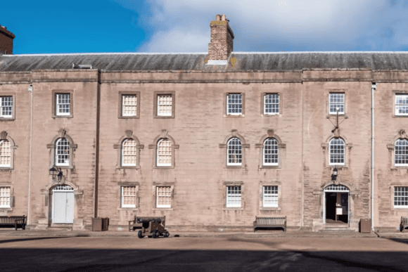 Front view of Berwick Barracks with Georgian-style stone buildings, arched windows, chimneys, and cannons in the courtyard.
